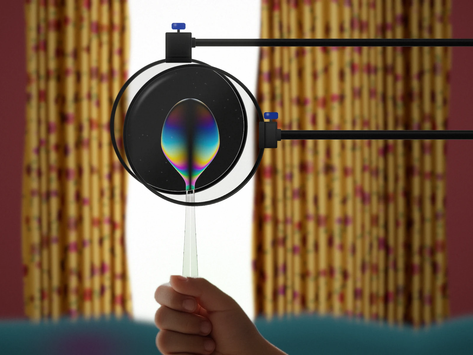 Two crossed polarizing filters held on black arms in front of a curtained window, with a clear plastic spoon between them. The spoon's bowl shows rainbow stress bands against the dark field, and a child's hand holds the handle below.